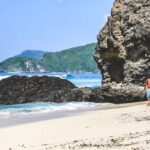 Lombok Solar - person in red shirt standing on seashore near brown rock formation during daytime