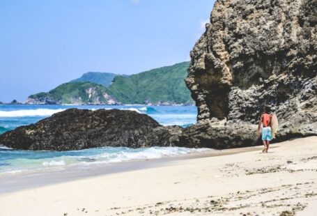 Lombok Solar - person in red shirt standing on seashore near brown rock formation during daytime