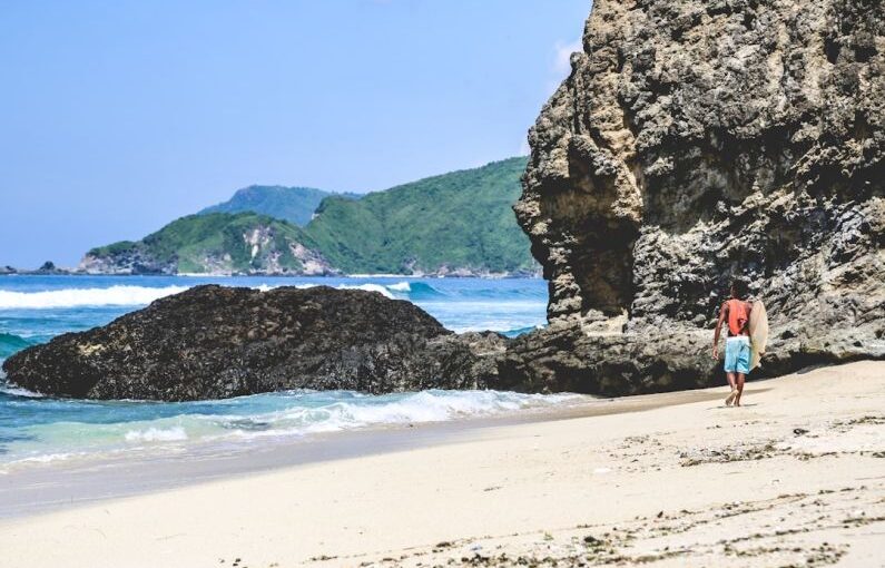 Lombok Solar - person in red shirt standing on seashore near brown rock formation during daytime