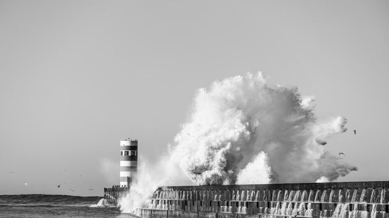 Coastal Resilience - a black and white photo of a large wave hitting a lighthouse